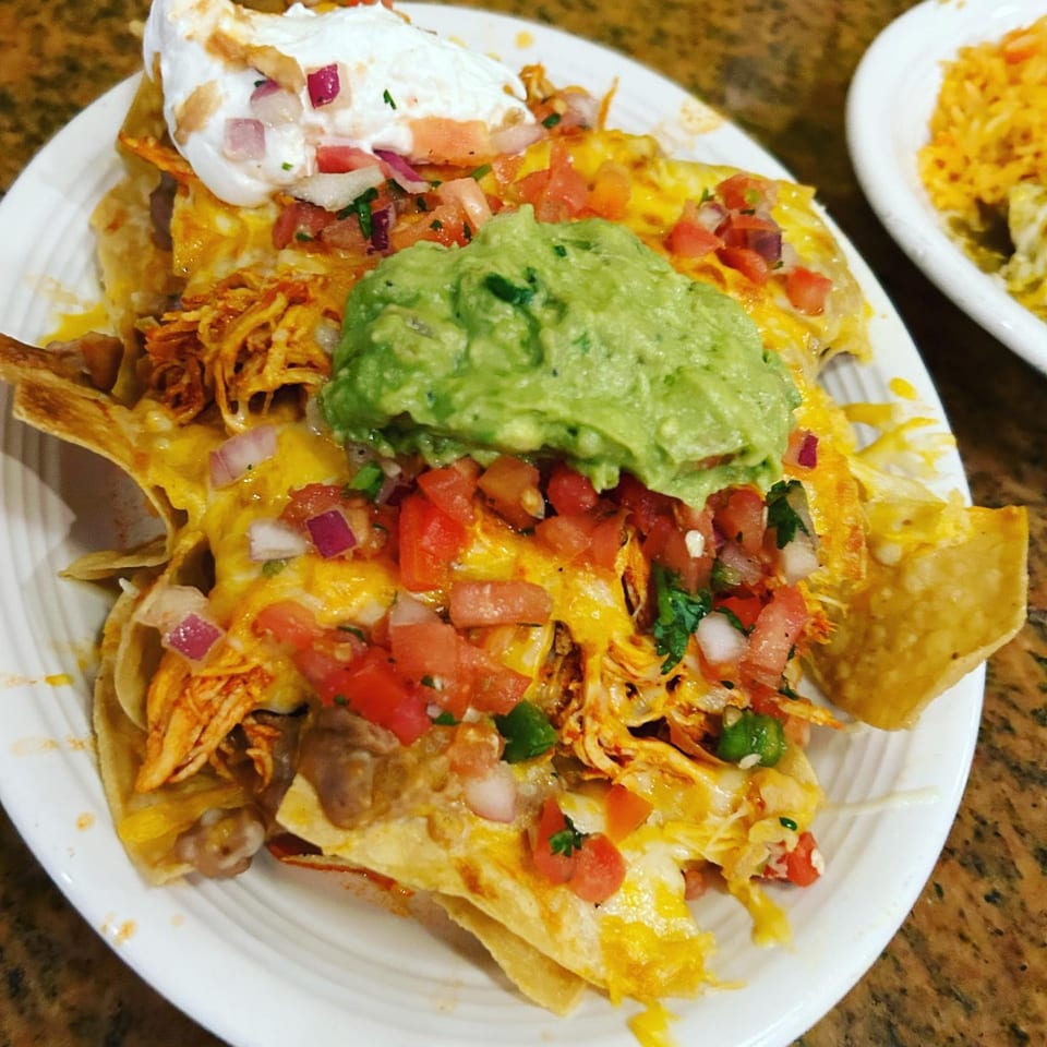 Chicken nachos with melted cheese, guacamole, pico de gallo, and sour cream on a white plate.