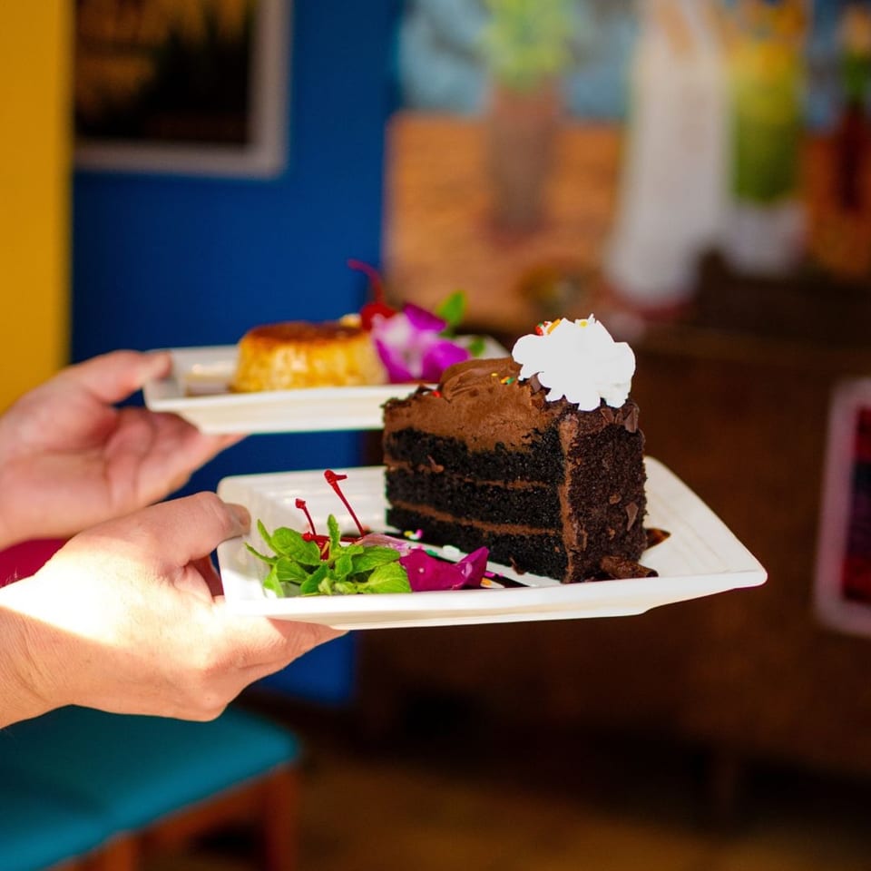 Hands holding plates with a slice of chocolate cake and a flan dessert.