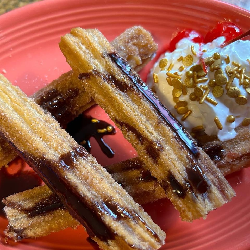 Churros drizzled with chocolate sauce, served with whipped cream, golden sprinkles, and a cherry on a red plate.